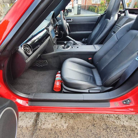 Mazda MX-5 Miata cabin with a red fire extinguisher mounted on a KAP bracket at the base of the left front seat
