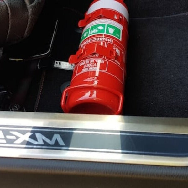 Close-up of a red fire extinguisher held in a KAP bracket at the base of the left front seat in a Mazda MX-5 Miata