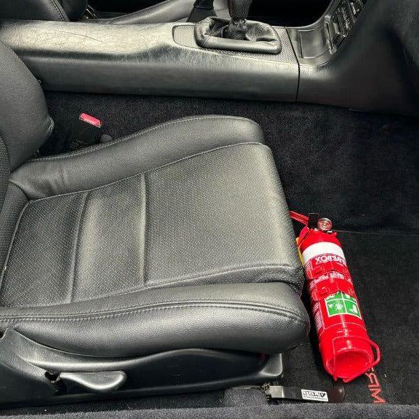 Close-up of a red fire extinguisher held in a KAP bracket at the base of the front seat in a Toyota MR2