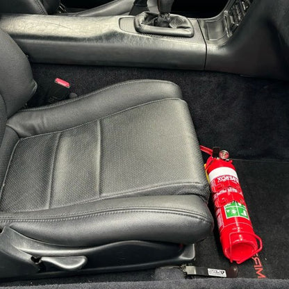 Close-up of a red fire extinguisher held in a KAP bracket at the base of the front seat in a Toyota MR2