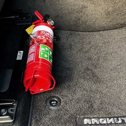Close-up of a red fire extinguisher held in a KAP bracket at the base of the front seat in a Toyota Land Cruiser