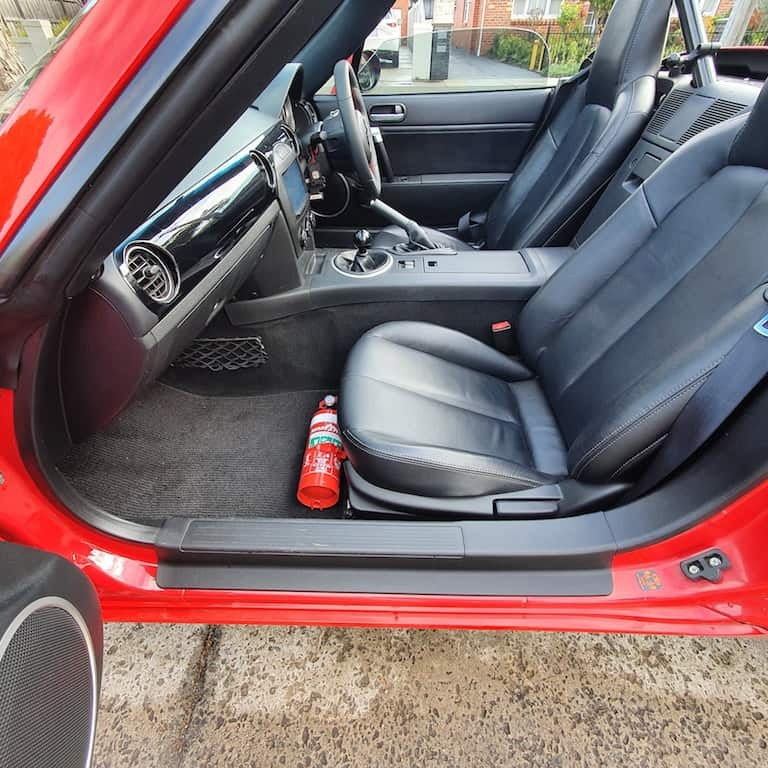 Mazda MX-5 Miata cabin with a red fire extinguisher mounted on a KAP bracket at the base of the left front seat