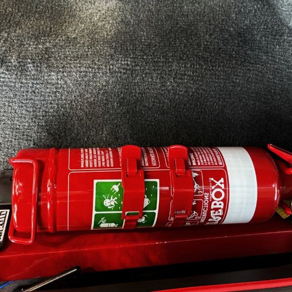 Close-up of a red fire extinguisher held in a KAP bracket at the base of the front seat in a Suzuki Samurai