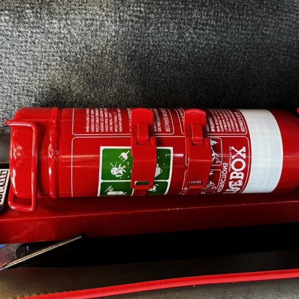 Close-up of a red fire extinguisher held in a KAP bracket at the base of the front seat in a Suzuki Samurai